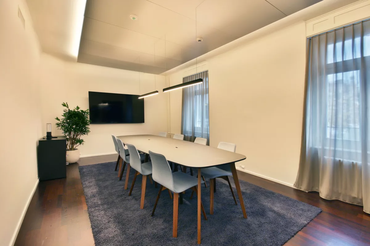 Moderner Konferenzraum, zentraler Holztisch mit acht Stühlen, TV, graue Vorhänge bei zwei Fenster, zwei dünne und lange Hängeleuchten über dem Tisch.