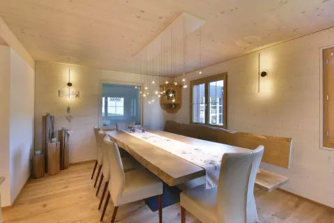 Essbereich mit Holzesstisch und fünf Stühle und eine Holzbank, Lampe mit Spotregen über dem Esstisch, Boden, Wände und Decke aus Holz, Lampen an den Wänden mit indirektem Licht