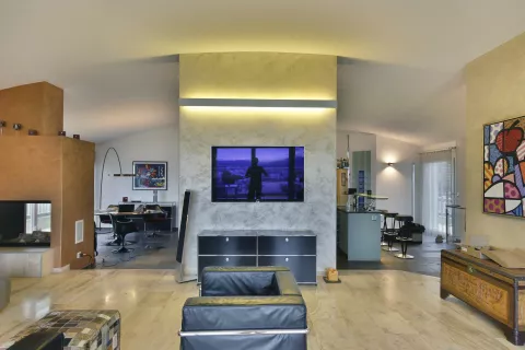 Wohnbereich mit an der Wand hängendem Fernseher, LED-Linie über dem Fernseher, offene Küche und Esszimmer im Hintergrund, Kamin, Sessel, Kunstbild an der Wand