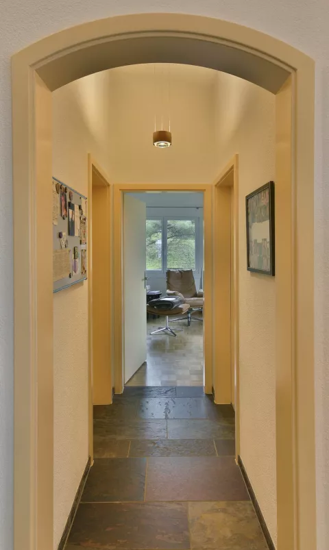 Flur mit Steinboden, runde Designerlampe, die von der Wand hängt, gelbe Wände, Türen rechts und links, offener Raum gegenüber mit Schreibtisch mit Sessel