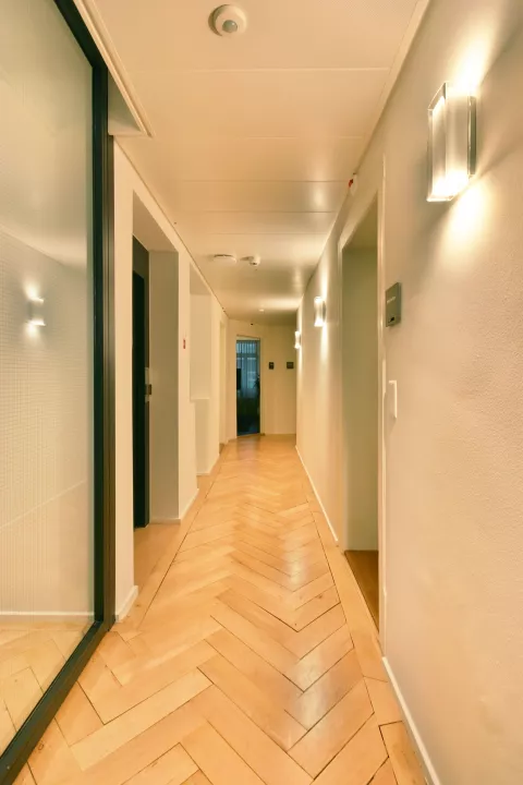 Korridor mit Parkettboden, Türen links und rechts entlang des Korridors, mehrere quadratische Lampen an den Wänden zwischen den diversen Türen
