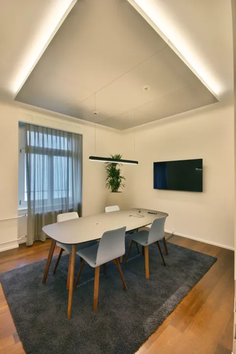 Moderner Konferenzraum, zentraler Holztisch mit vier Stühlen, TV, graue Vorhänge beim Fenster, dünne und lange Hängeleuchten über dem Tisch.