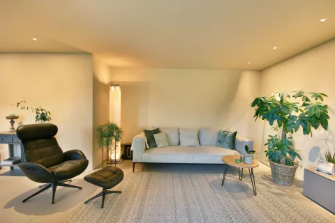 Wohnbereich mit schwarzen Sessel, graues Sofa, kleiner Couchtisch, grüne Pflanzen, LED-Spots in der Decke und hohe helle Stehlampe in der Ecke